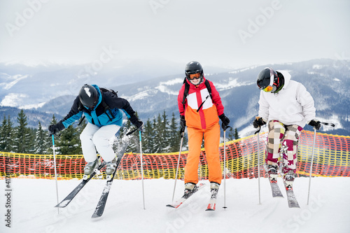 Full length of skiers team in ski jackets and helmets skiing in winter snowy mountains, making jump while sliding down snow-covered slope on skis. Concept of winter sport activities and friendship.