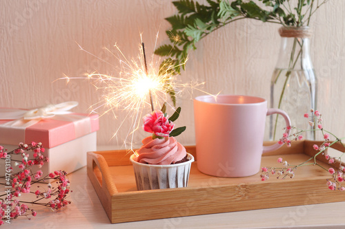 Party time. Birthday cupcake with sparkler, cup of coffee on wooden tray, gift box and eucalyptus leaf decor. Close-up