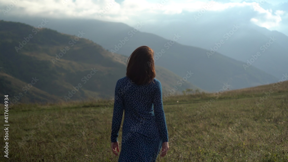 A young woman walks in a blue dress and raises her hands up, looking at the mountains.