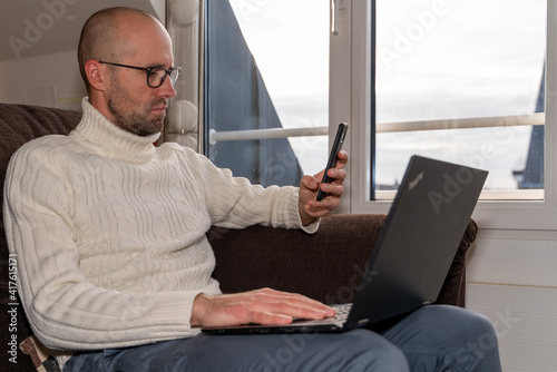 homme adulte assis dans un canapé en train de travailler sur son ordinateur et regardant son téléphone portable, travail à la maison, à distance