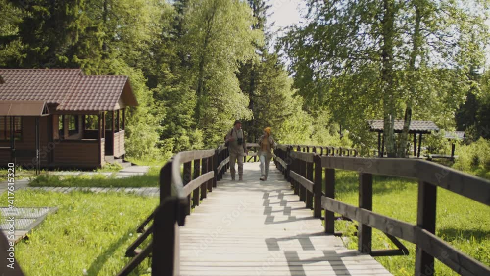 Wide shot of senior tourists walking towards the camera on wooden stairs in park and talking while hiking together on summer day Wide shot of senior tourists walking towards the camera on wooden stairs in park and talking while hiking together on summer day