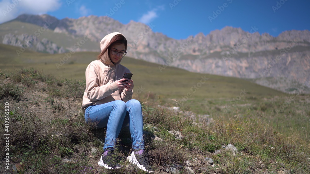 A young woman in a sweatshirt sits with a phone in her hands against a background of mountains.