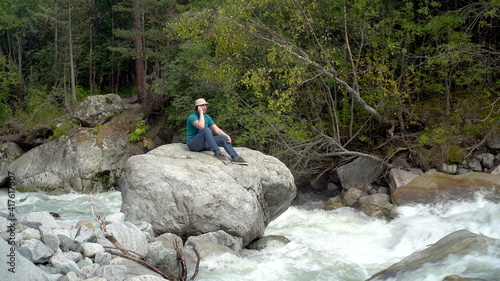 A young man in a Panama hat sits on a stone and speaks on the phone in the mountains by the river. Travel to the mountains.