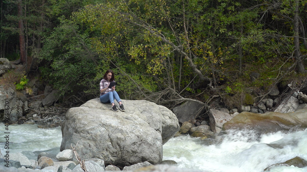 A young woman with glasses sits on a stone with a tablet in his hands in the mountains near the river. Travel to the mountains.
