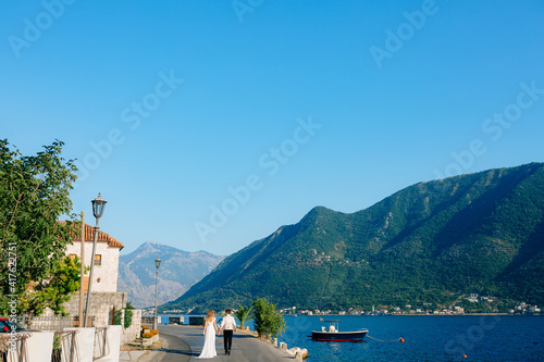 The bride and groom walk holding hands on the pier in Perast against the backdrop of a picturesque view of the Bay of Kotor 