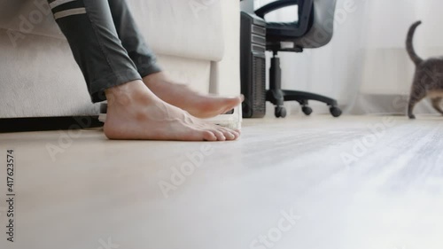 The robot vacuum cleaner moves around the room. The woman raises her legs so that the vacuum cleaner passes. The cat is in the background