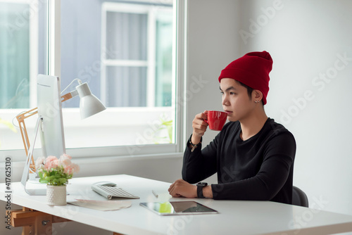 Young designer asian man drinking coffee, analyzing data looking at computer screen at office.