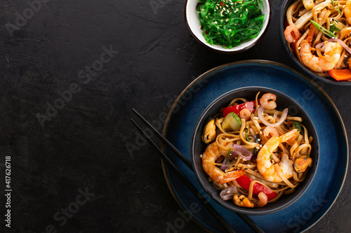 Fried noodles or wok noodles with shrimps, vegetables and mussels in a black bowl and seaweed salad on a black textured background, top view, copy space. Traditional Asian food.