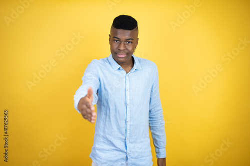 Young african american man wearing a casual shirt standing over yellow background smiling friendly offering handshake as greeting and welcoming