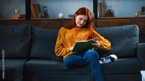 Smiling red haired girl reading book on sofa