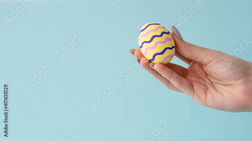 Easter ornament. Holiday decoration. Holiday symbol. Greeting card. Handmade yellow color egg with pink wave stripe design in female hand isolated on blue copy space background.