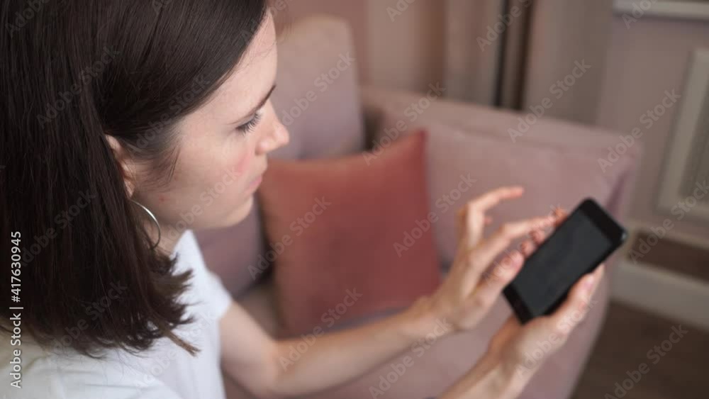 Relaxed young woman using smartphone browsing social networks, playing mobile games or texting while sitting on the couch. Millennial woman spends time at home with mobile gadgets. Side view Relaxed young woman using smartphone browsing social networks, playing mobile games or texting while sitting on the couch. Millennial woman spends time at home with mobile gadgets. Side view