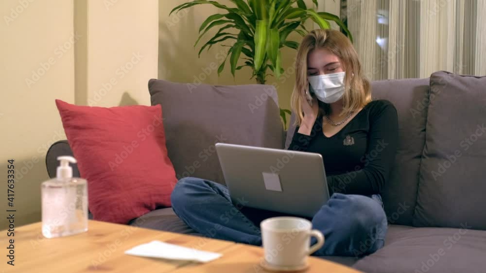 Young Turkish woman with a facial mask, using computer and talking with her smartphone while sitting on the sofa. There is a coffee cup, hand sanitizer and a mask on the table.