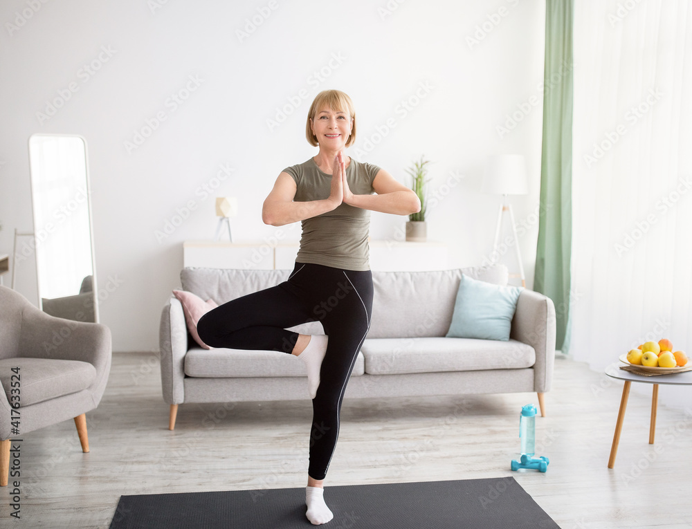 Fototapeta premium Domestic yoga practice. Positive mature lady standing in tree pose, keeping balanced during covid lockdown at home