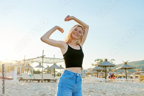 Beautiful happy female teenager on sandy beach with hands raised up dancing at sunset
