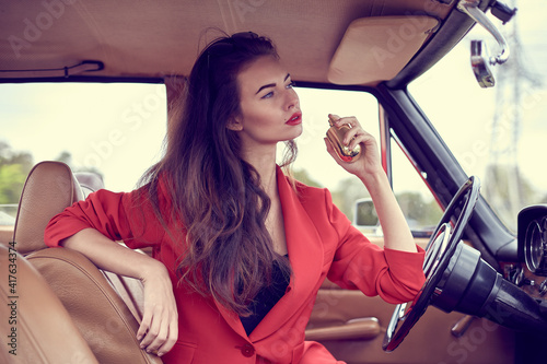 Beautiful young vogue woman in red suit sitting in retro car using a perfume. photo