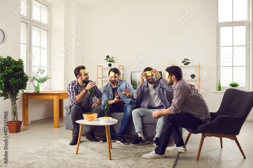 Men's leisure. Group of male friends sit on a sofa in the living room with stickers on their foreheads and play the game Who am I. Friends have fun at home playing games and drinking beer with chips.