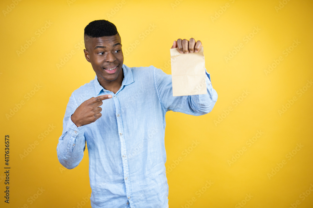 Young african american man wearing a casual shirt standing over yellow background smiling, surprised and pointing paper bag Young african american man wearing a casual shirt standing over yellow background smiling, surprised and pointing paper bag