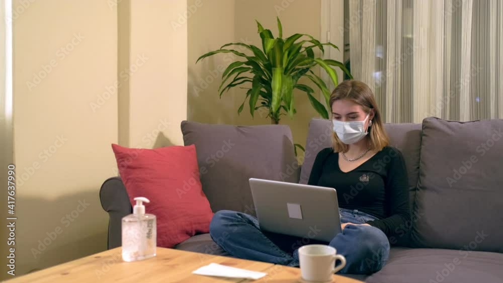 Young Turkish woman with a facial mask, having a video call on her computer while sitting on the sofa. There is a coffee cup, hand sanitizer and a mask on the table. Young Turkish woman with a facial mask, having a video call on her computer while sitting on the sofa. There is a coffee cup, hand sanitizer and a mask on the table.