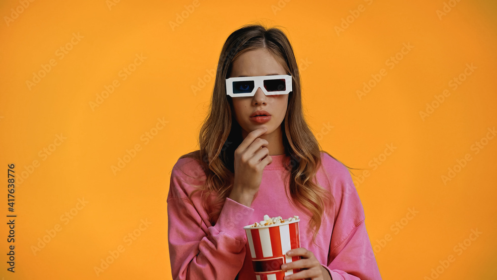 Obraz premium teenage girl in 3d glasses holding bucket and eating popcorn isolated on yellow