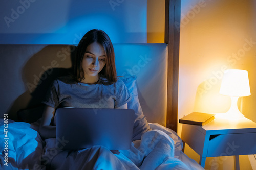 Internet night. Home leisure. Digital communication. Modern technology. Relaxed tired woman enjoying loneliness with laptop in bed late in dark bedroom with blue warm yellow lamp light.