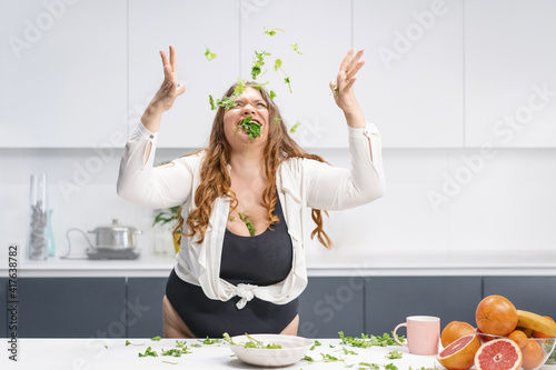 Happy chubby girl scattering spinach leaves all over the table. Overweight girl happy loosing weight eating fresh salad. Curvy body young woman with long blond hair. Dieting and nutrition concept.  photo