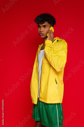curly african american man in jacket and shorts talking on smartphone isolated on red © LIGHTFIELD STUDIOS