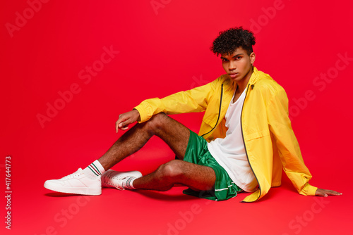 full length of pierced african american man in yellow jacket and white sneakers sitting while posing on red