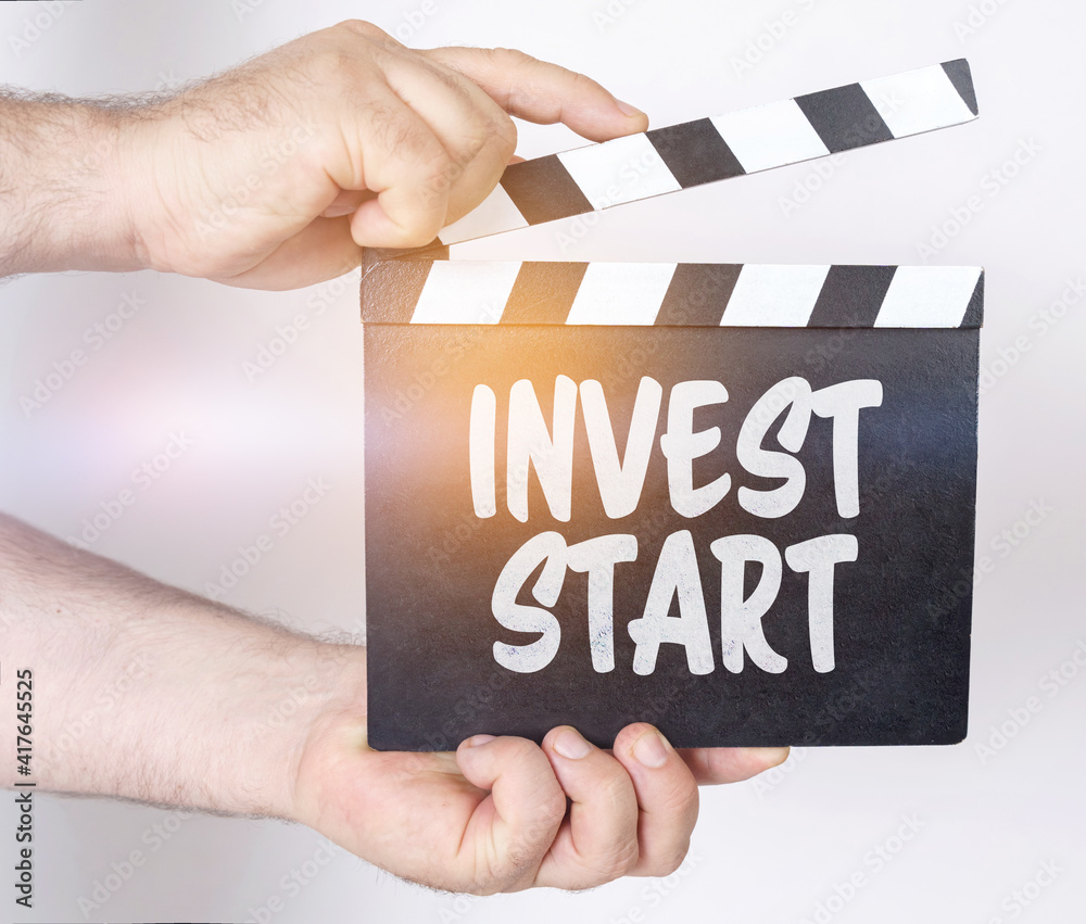 Obraz premium On a white background, a man holds a clapperboard in his hands on which it is written - INVEST START