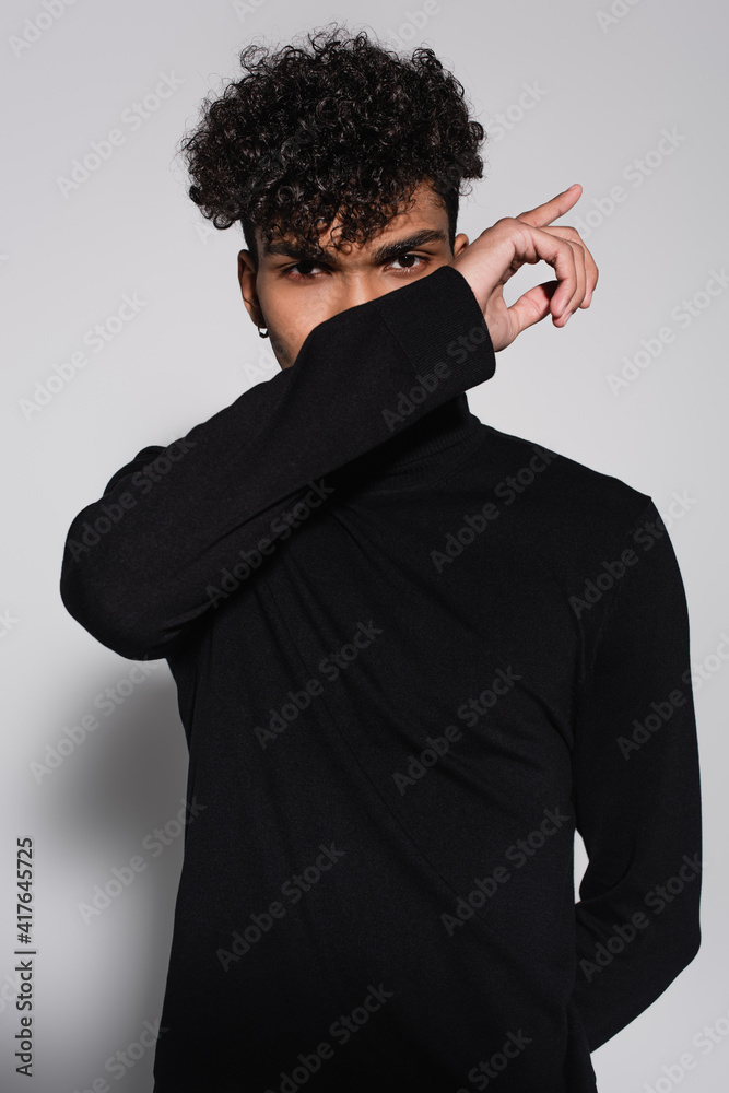 curly african american man covering face with hand while posing on grey