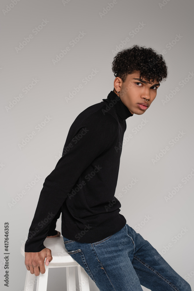 african american young man in turtleneck sweater leaning on white chair isolated on grey african american young man in turtleneck sweater leaning on white chair isolated on grey