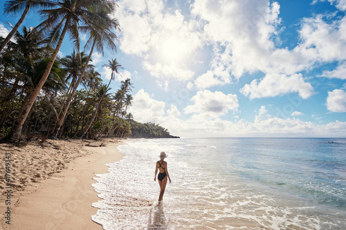 Vacation on the seashore. Back view of young woman walking away on the beautiful tropical white sand beach.