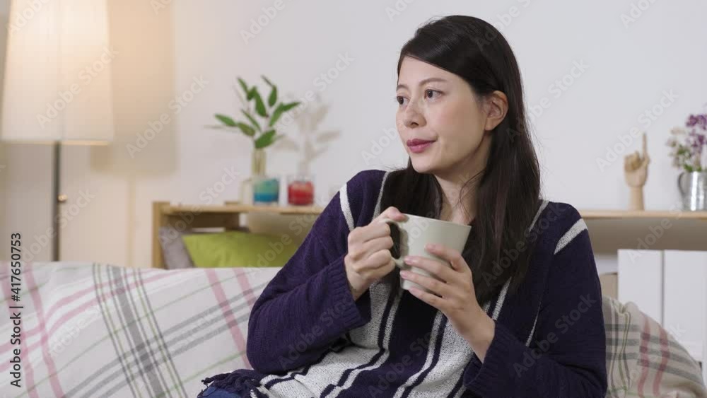 portrait charming chinese woman seated on home sofa is sipping tea and tucking her hair while gazing afar in contemplation. portrait charming chinese woman seated on home sofa is sipping tea and tucking her hair while gazing afar in contemplation.
