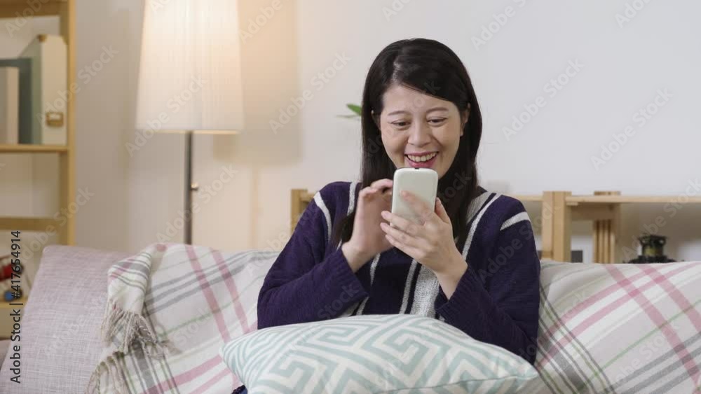 pretty asian girl is laughing happily and looking at screen with her eyes glowing in surprise while texting with her friend on the phone in the living room. pretty asian girl is laughing happily and looking at screen with her eyes glowing in surprise while texting with her friend on the phone in the living room.