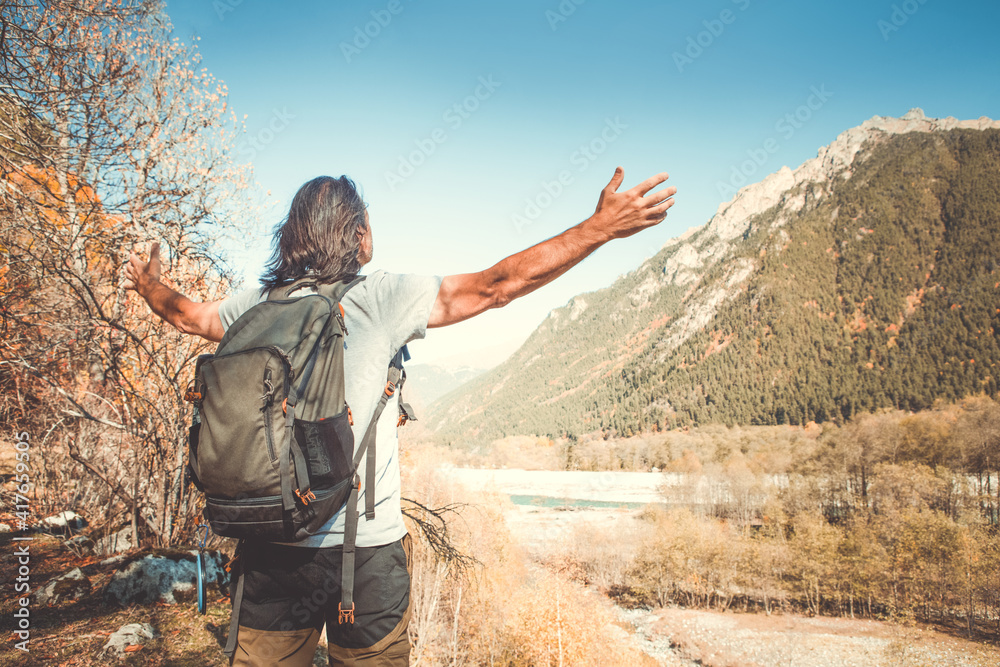 A man with a backpack against the background of blue sky and mountains. He spread his arms wide and looks ahead. Autumn. back view.