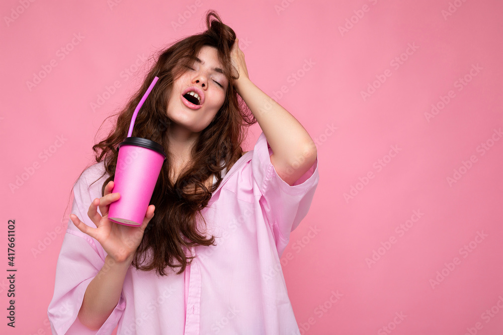 Fototapeta premium Shot of beautiful sexy young positive brunette curly woman wearing pink shirt isolated over pink background holding paper coffee cup for mockup drinking and enjoying