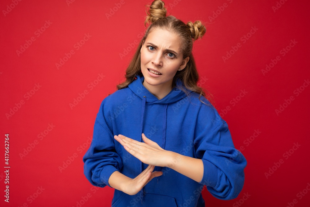 Photo of emotional young attractive blonde woman with two horns with sincere emotions wearing hipster bright blue hoodie isolated over red background with copy space and showing pause sign Photo of emotional young attractive blonde woman with two horns with sincere emotions wearing hipster bright blue hoodie isolated over red background with copy space and showing pause sign