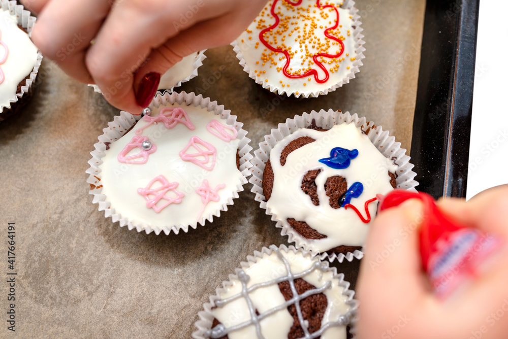 Fototapeta premium A woman squeezes colored frosting from a tube onto chocolate brown cupcakes covered with white frosting with colorful decorations.
