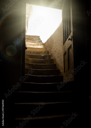 Old wooden door open into basement with light rays shining into dark creepy cellar from stone worn staircase, rundown with shadows and darkness.