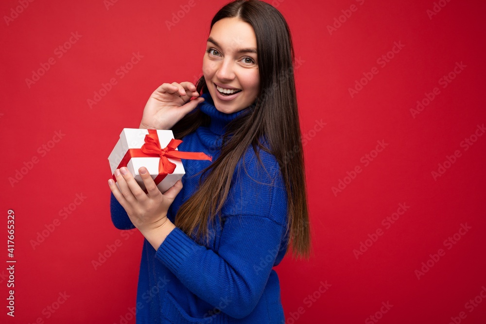 Fototapeta premium Shot of beautiful positive smiling young brunette woman isolated over red background wall wearing blue casual sweater holding white gift box with red ribbon and looking at camera