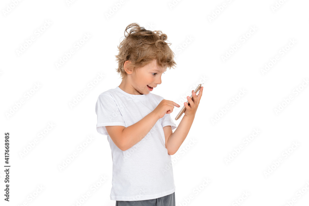 Cute Caucasian kid boy in a white T-shirt on a white isolated background stands with a cell phone in his hand, uses a smartphone Cute Caucasian kid boy in a white T-shirt on a white isolated background stands with a cell phone in his hand, uses a smartphone