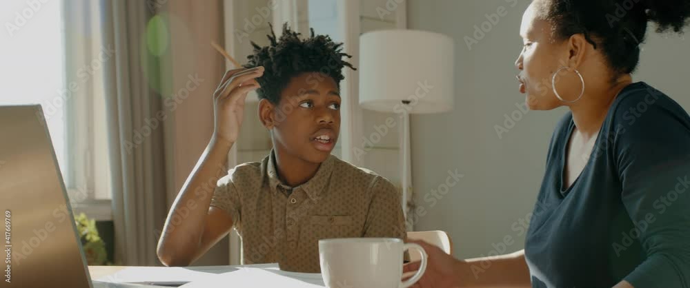 African American Black mother sits near her son, helping him with homework. Distance learning from home during COVID-19 coronavirus pandemic, stay home concept. Shot with 2x anamorphic lens