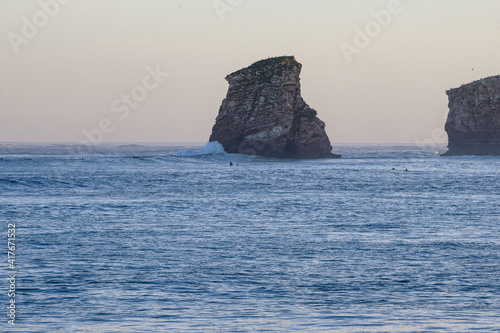 the twins, rocks of the beach of Hendaye at dawn surrounded by waves and in the distance people surfing. The sky behind the rocks is orange by the sunrise sun and strong waves breaking 