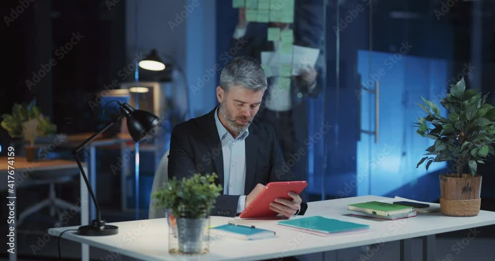 Businessman adult in formal suit sitting by desk using tablet computer management application working late night in corporate coworking space. Businessman adult in formal suit sitting by desk using tablet computer management application working late night in corporate coworking space.