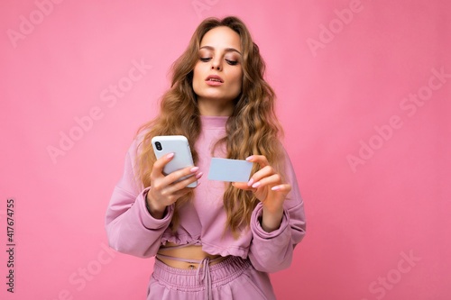 Beautiful young blonde curly woman wearing pink clothes isolated over pink background using mobile phone paying online shopping through credit card looking at smartphone display
