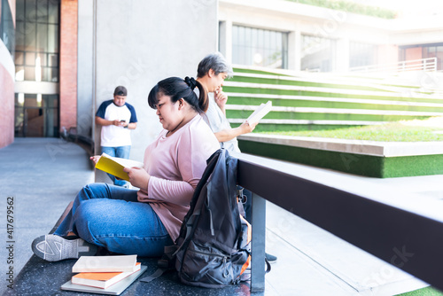 Asian woman student, sitting and reading a book To review the lesson, with blur of two man which her friends standing reading and use a mobile phone background, to people and adult education concept.