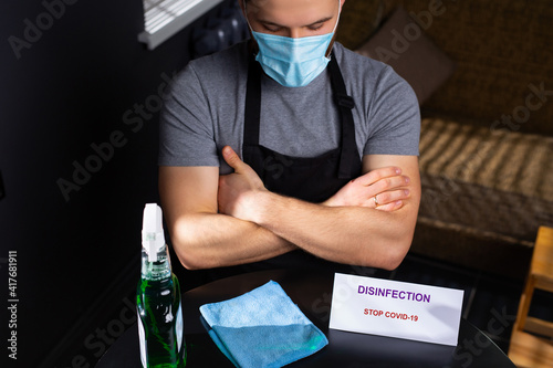 Disinfection during covid 19. Photo from above of a man in an apron and a medical mask. There is a sign and disinfectant on the table.