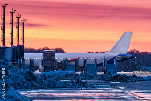 A passenger jet plane stands on the airport apron covered with snowdrifts near a row of aircraft mobile gangway against the backdrop of a picturesque crimson sunset photo