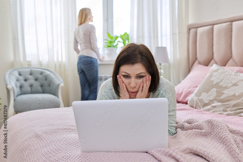 Surprised mature woman with laptop at home in bed, woman near window background Surprised mature woman with laptop at home in bed, woman near window background