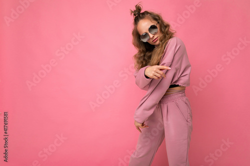 Attractive joyful young blonde woman wearing everyday stylish clothes and modern sunglasses isolated on colorful background wall looking at camera